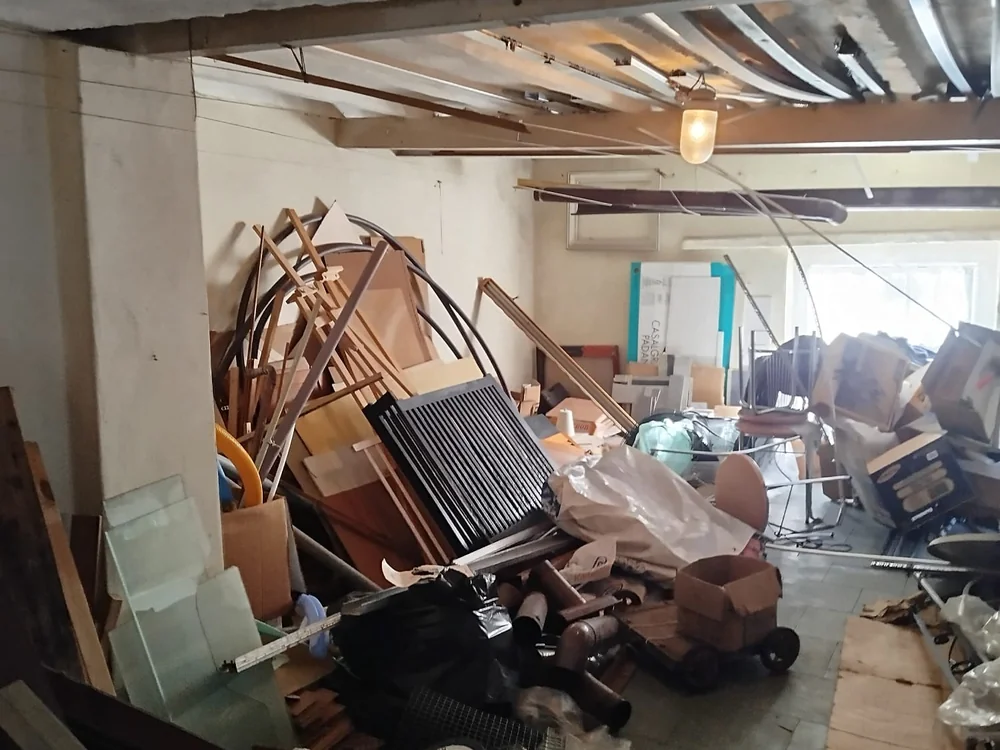 Piwnica przed opróżnieniem: stos metalowych rur, kartonów i sprzętu pod sufitem z instalacją